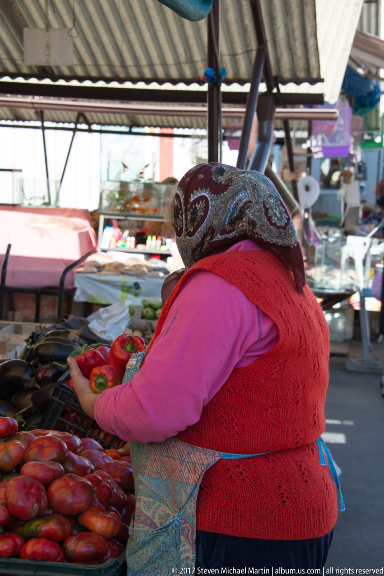 Headscarfed market vendor in a red vest leaning over and sorting a large display of ripe tomatoes.