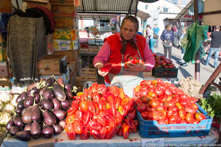 Vendor behind a market display of eggplants, red peppers, tomatoes, garlic, and other fresh vegetables.