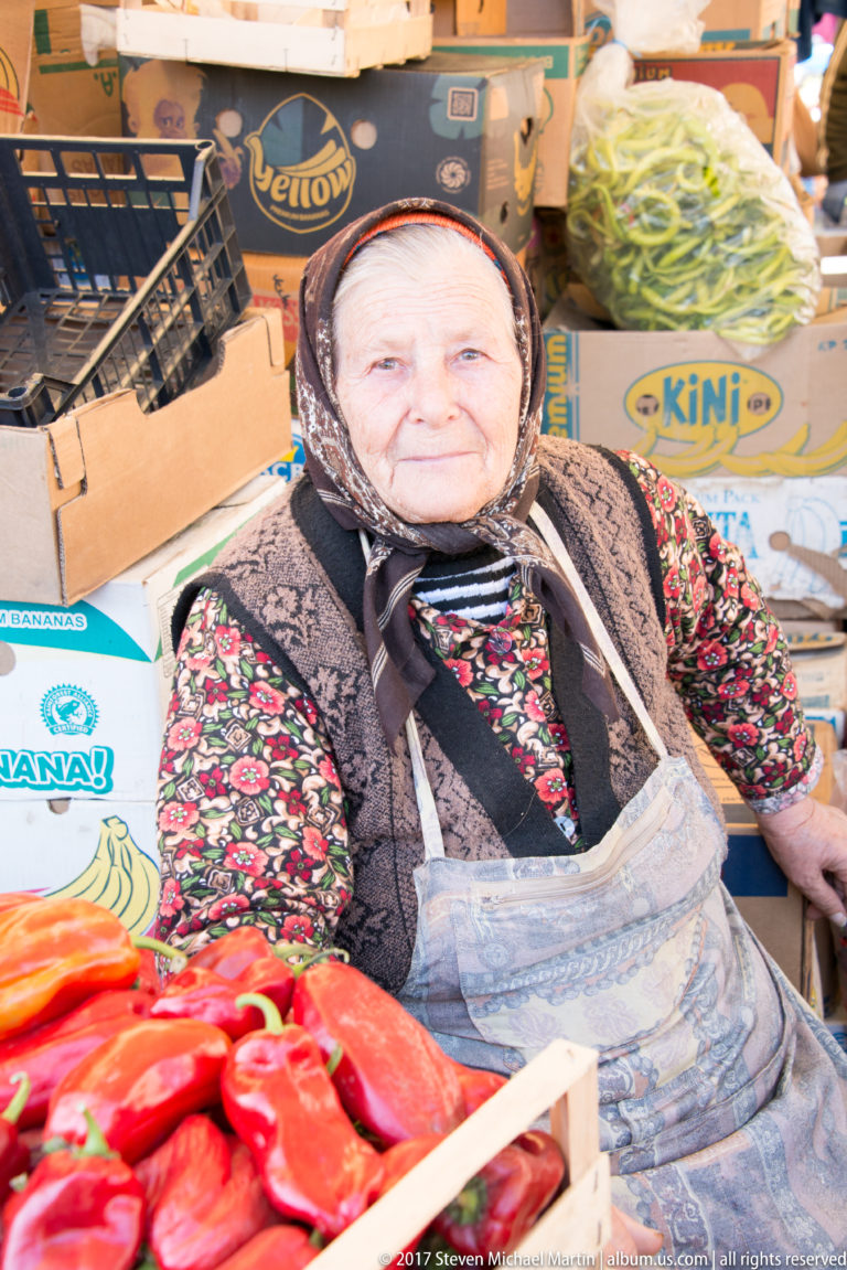 Elderly market seller in patterned headscarf, floral blouse, vest, and apron seated behind bright red peppers.