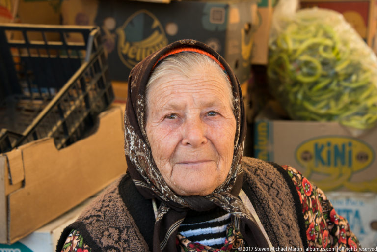 Close portrait of an elderly market seller in patterned headscarf with produce boxes and green peppers behind her.