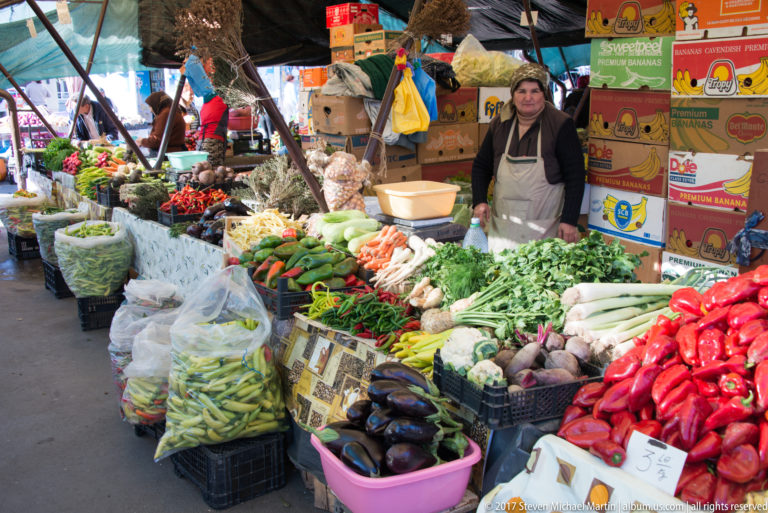 Wide market stall piled with peppers, eggplants, carrots, greens, beans, root vegetables, sacks, and a standing vendor.