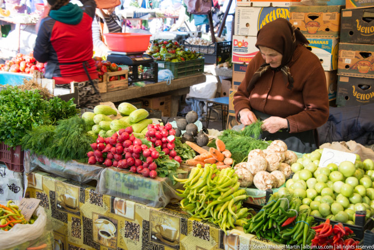 Market vendor preparing leafy greens behind displays of radishes, carrots, peppers, root vegetables, and onions.