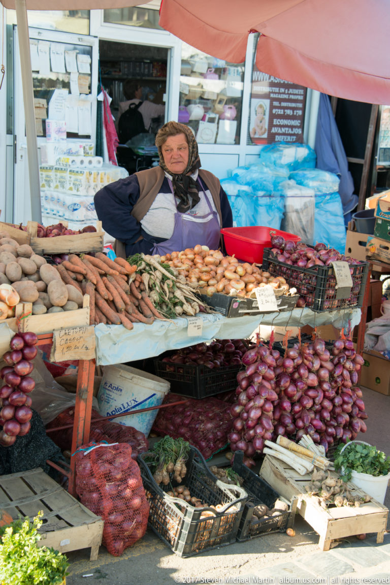 Seated vendor behind a market stall filled with red onions, root vegetables, garlic, bundles, crates, and price signs.
