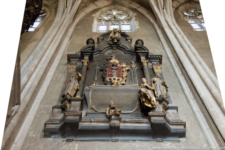 Ornate wall-mounted church epitaph with heraldic crest, inscription panel, sculpted figures, and Gothic wall ribs.