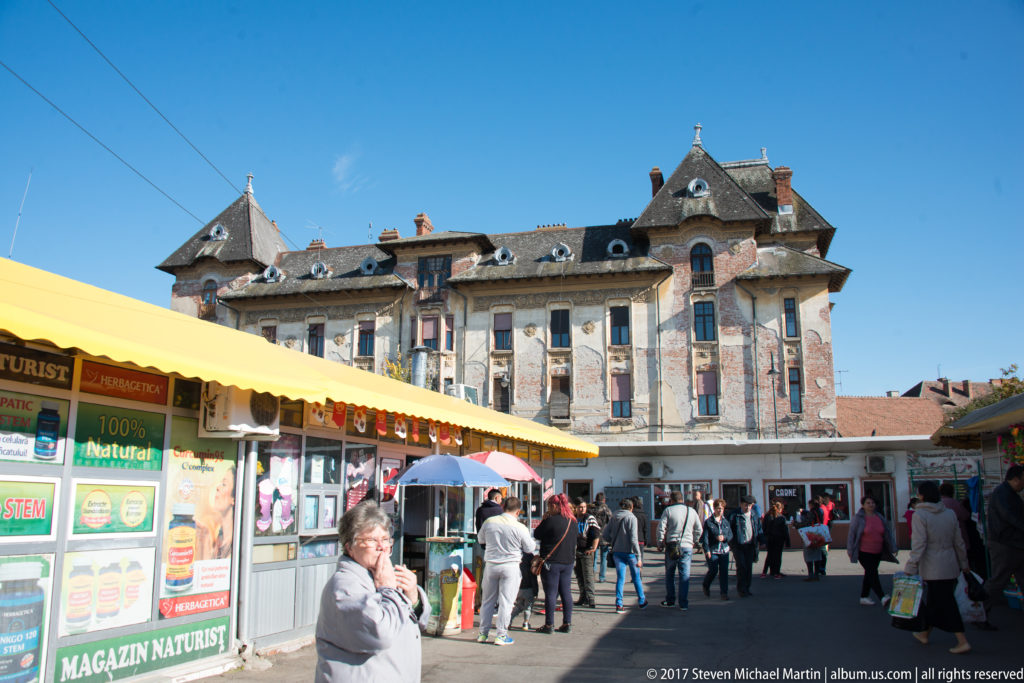 Open-air Sibiu market with shoppers, stalls, umbrellas, and an ornate historic building rising behind the courtyard.