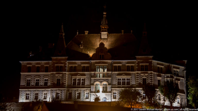 Large ornate historic public building illuminated at night, with tall windows, arched entrance, steep roof, and central turret.