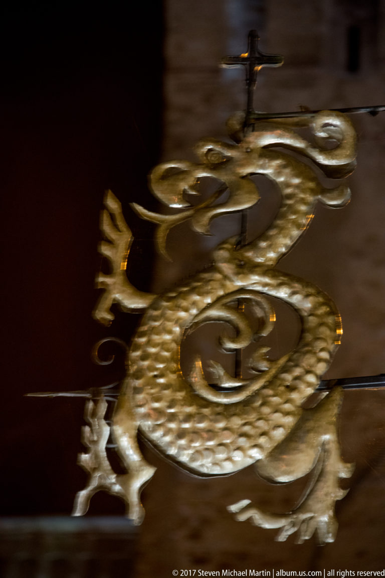 Coiled dragon-shaped metal ornament with open mouth, curled tail, and projecting limbs against a dark background.
