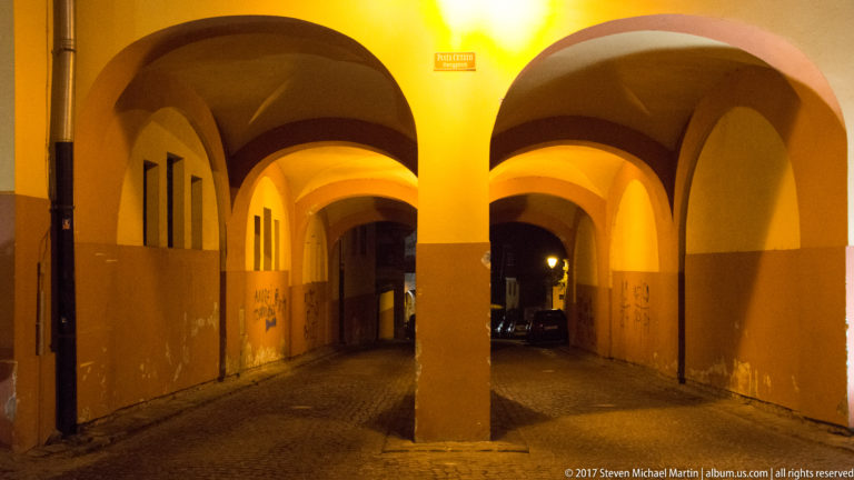 Yellow-lit vaulted passage divided by a central pier, with two arched routes opening toward a dark street.