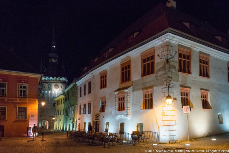 Illuminated pale historic buildings, café tables, pedestrians, and cobbled paving in Sighisoara's citadel at night.