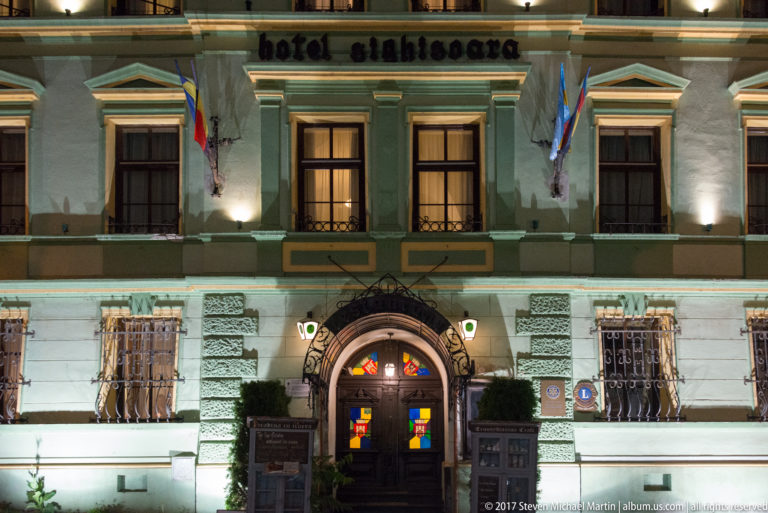Illuminated green Hotel Sighisoara facade with heraldic arched entrance, flags, and barred lower windows.