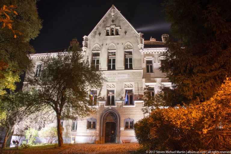 Illuminated Joseph Haltrich school facade with steep central gable, arched entrance, tall windows, flags, and trees.