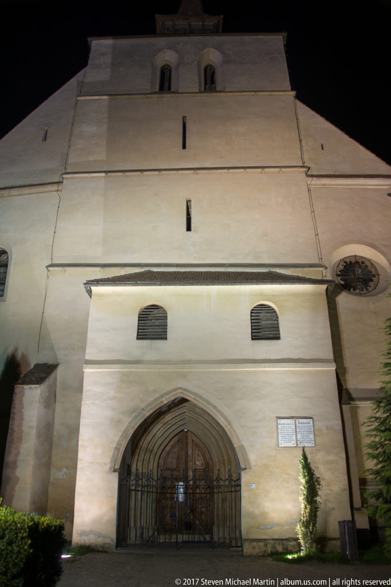Large pale church facade at night with pointed-arch wooden doorway, narrow openings, and a circular window.