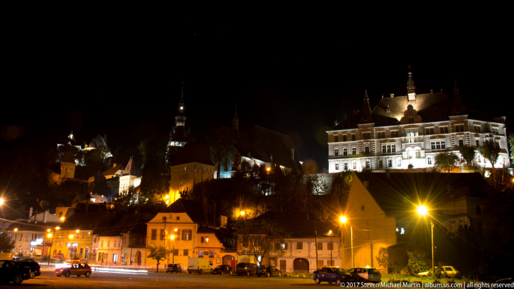 Wide night panorama of Sighisoara's illuminated citadel hill, towers, historic buildings, and lower-town streetlights.