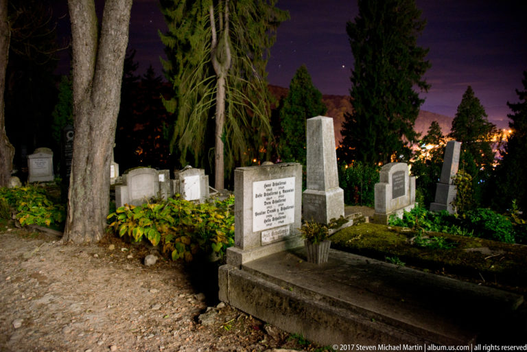 Inscribed rectangular grave and headstone surrounded by older monuments, plants, and large trees at night.