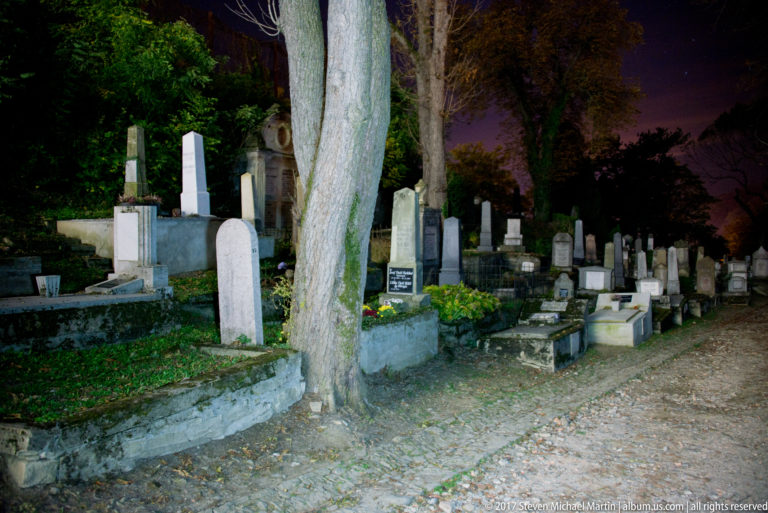 Rows of old graves and stone monuments beneath large trees along an unpaved cemetery path at night.