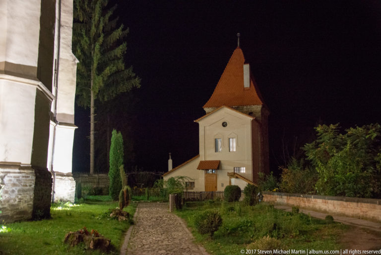 Small pale tower with steep red-tiled roof beside a cobbled path, shrubs, and tall evergreens at night.