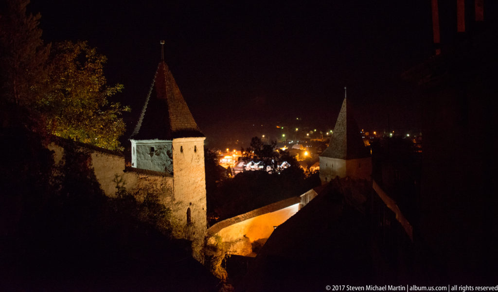 Two steep-roofed stone defensive towers connected by an illuminated wall above the lights of Sighisoara at night.