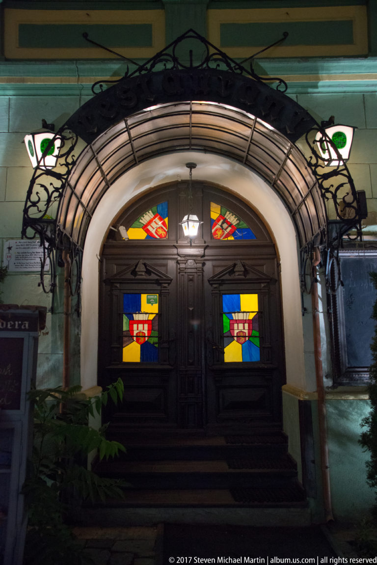 Arched dark-wood entrance with colorful heraldic glass panels and decorative metalwork illuminated at night.