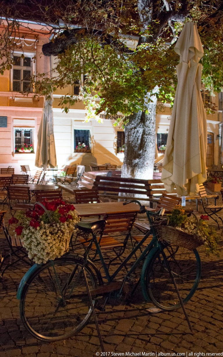 Bicycle leaning among empty café tables, closed parasols, flowers, and a large tree on a cobbled terrace at night.
