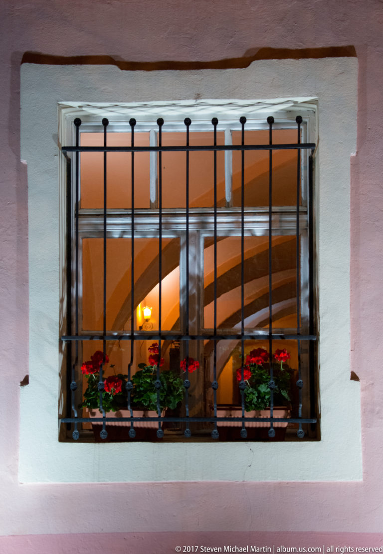 Black iron bars cover a wooden window with red flower boxes in a pale plaster wall, warmly lit from inside.