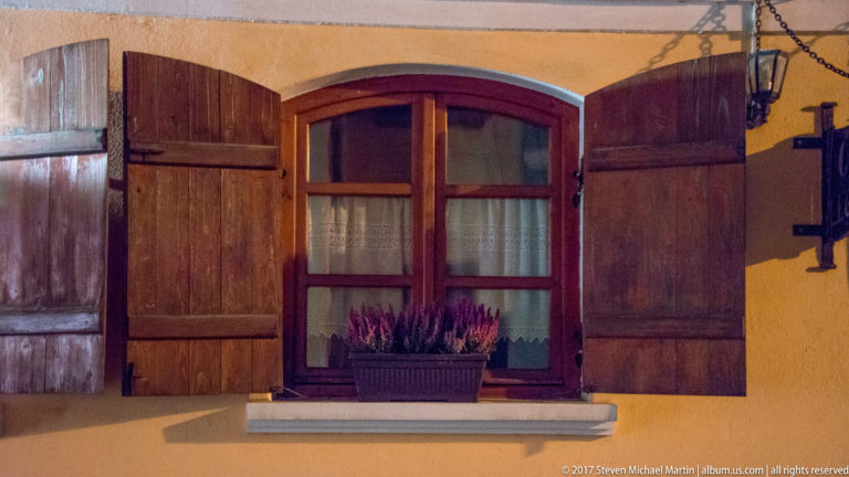 Red-brown wooden window with open shutters and a flower box set in a yellow plaster wall.