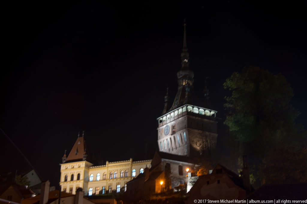 Sighisoara Clock Tower rising above illuminated historic buildings on the citadel hill at night.