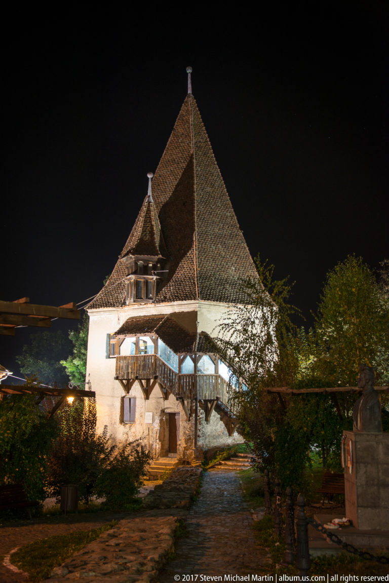 Historic house with a very steep tiled roof and exterior timber stair, illuminated at night.