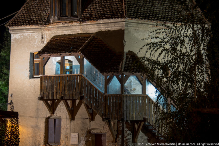 Covered wooden exterior stairway with railings and small tiled roofs attached to a pale historic building.