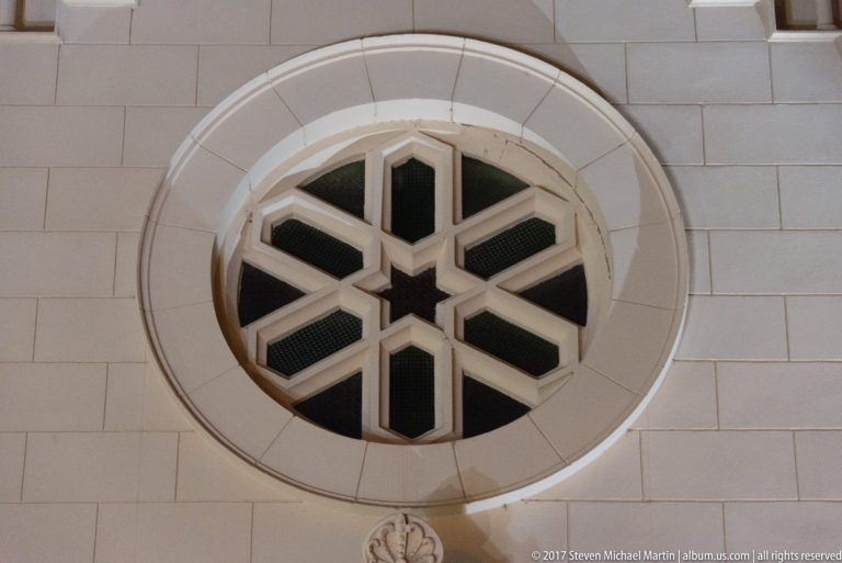 Circular radial window with white geometric mullions and dark glazing set in a pale masonry facade.
