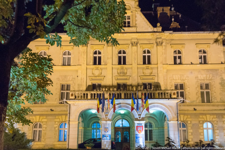 Wide night view of a symmetrical public building with arched entrance, balcony, flags, and shield emblems.