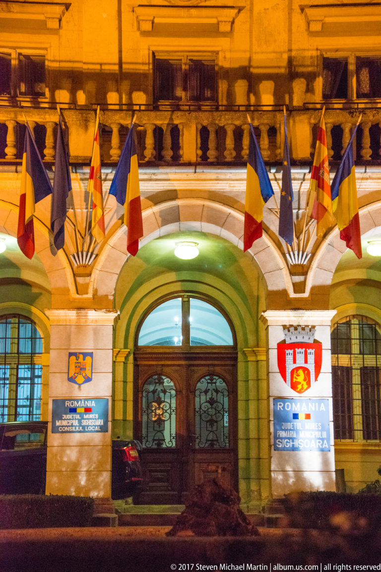 Illuminated arched civic entrance decorated with Romanian and European Union flags and shield emblems.