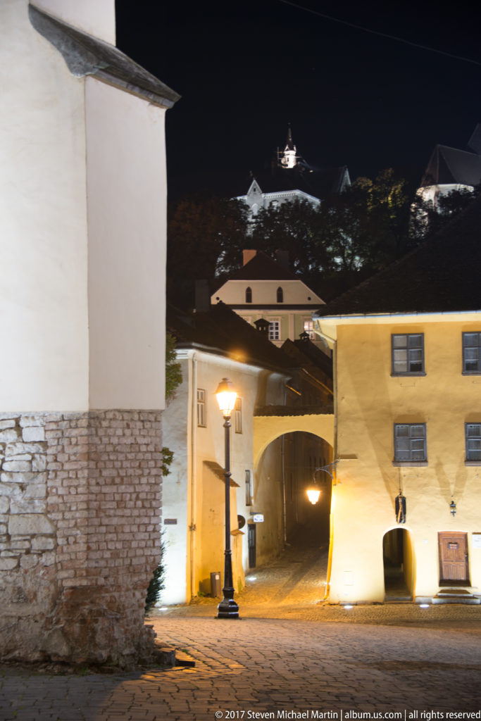 Illuminated arched passage and pale buildings below the dark hilltop citadel at night.