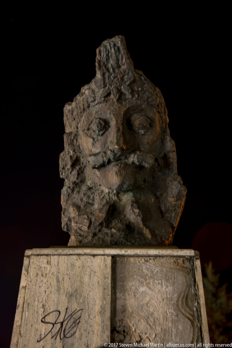 Close night view of rough abstract sculpture above a pale stone pedestal with graffiti.