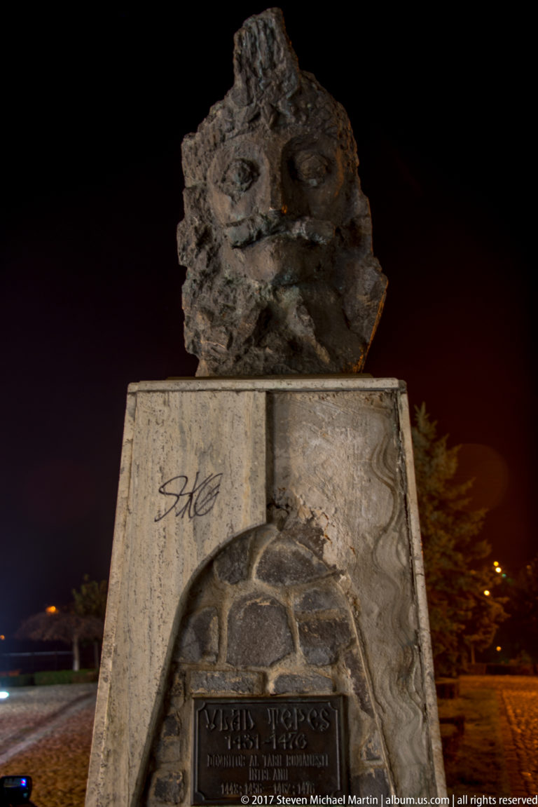 Dark abstract memorial sculpture on a stone pedestal with a Vlad Tepes plaque at night.