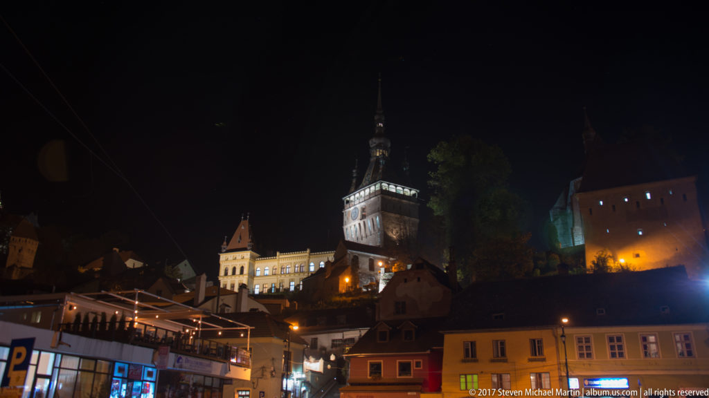Night cityscape with colorful lower-town buildings beneath Sighisoara's illuminated hilltop citadel and Clock Tower.