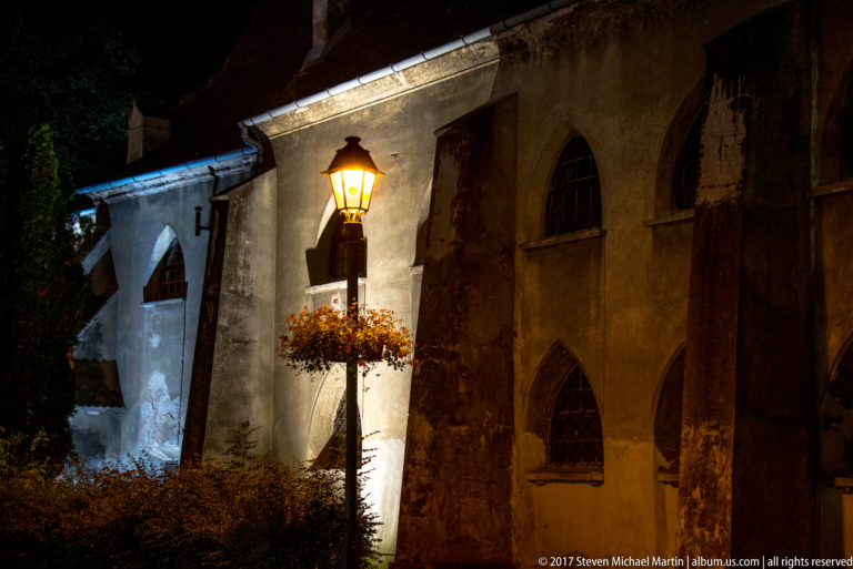 Night-lit church wall with pointed-arch windows, a recessed doorway, and a lamppost with flowers.