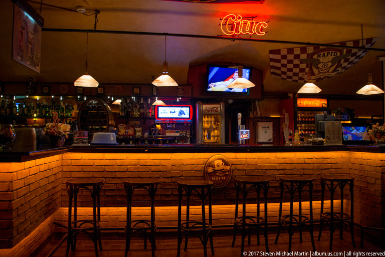 Brick-fronted bar counter with stools, pendant lamps, coolers, televisions, and sports decorations.
