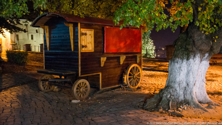 Dark wooden wheeled kiosk with colorful panels parked beside a mature tree on cobblestones at night.