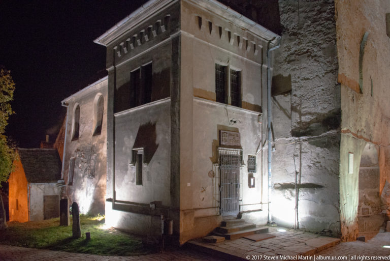 Small pale historic building with barred doorway beside a rough masonry wall at night.