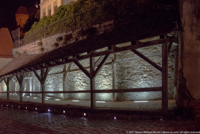 Timber posts and braces support a covered passage beside a lit rough-stone defensive wall.