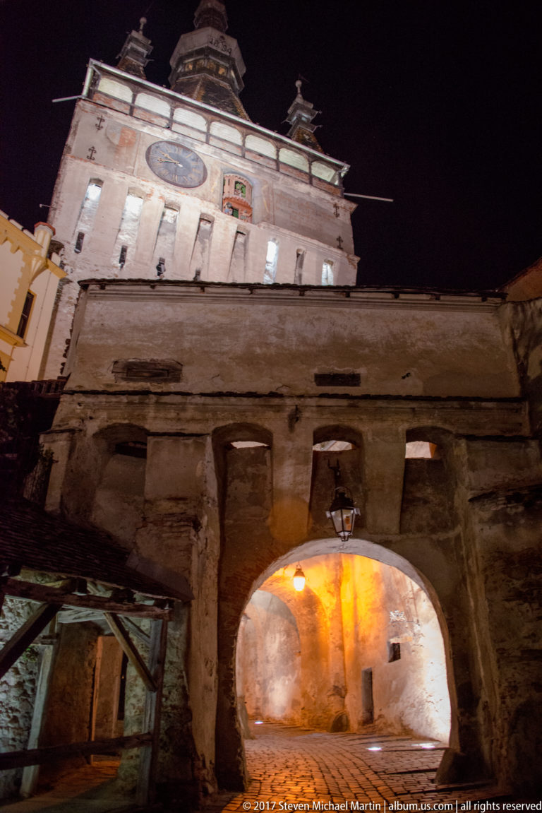 Large illuminated archway beneath Sighisoara Clock Tower, with weathered masonry and timber gallery elements.