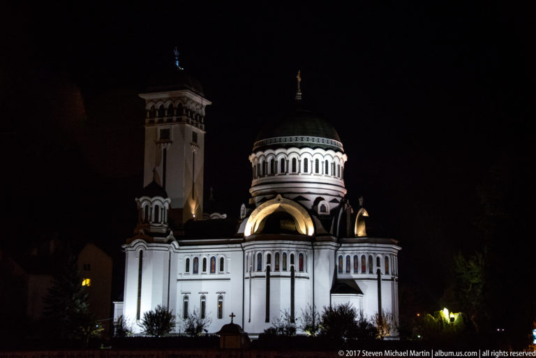 White illuminated Orthodox church with central dome, arched windows, and tall bell tower against the night sky.