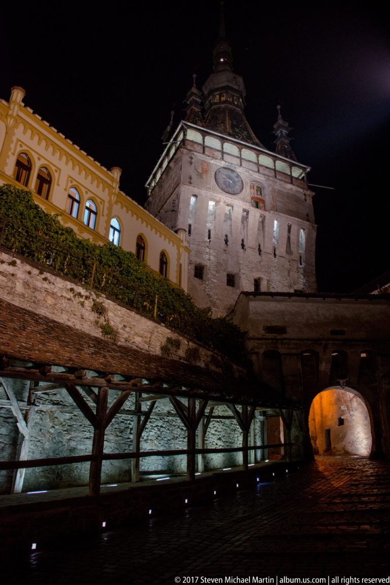 Sighisoara Clock Tower above a timber-supported wall walk and illuminated arched gateway at night.