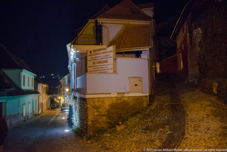 Forked cobbled lane at night between plastered and rough-stone buildings in Sighisoara's citadel.