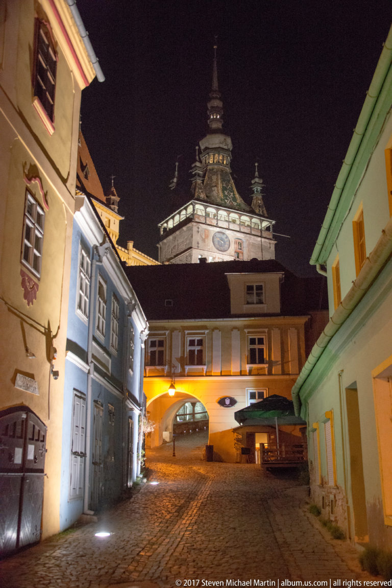 Narrow cobbled street and colorful historic houses framing Sighisoara Clock Tower at night.