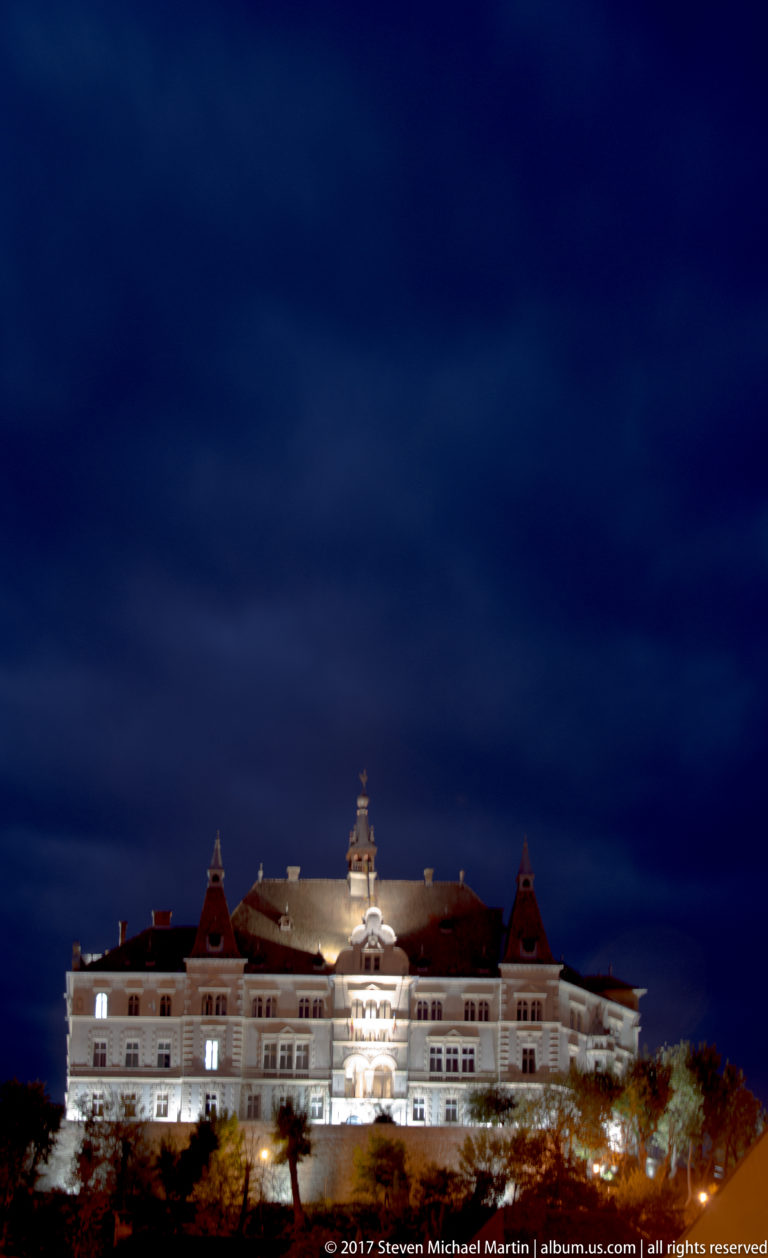 Wide night view of a large illuminated historic building partly screened by trees on a slope.