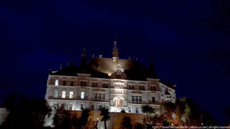 Large illuminated historic building with many windows, steep roofs, and a central vertical feature at night.