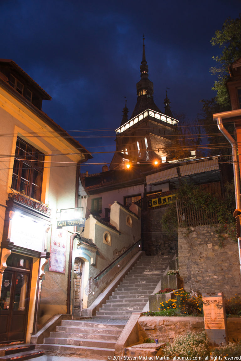 Stone steps rise between buildings toward the illuminated upper stage of Sighisoara Clock Tower at night.