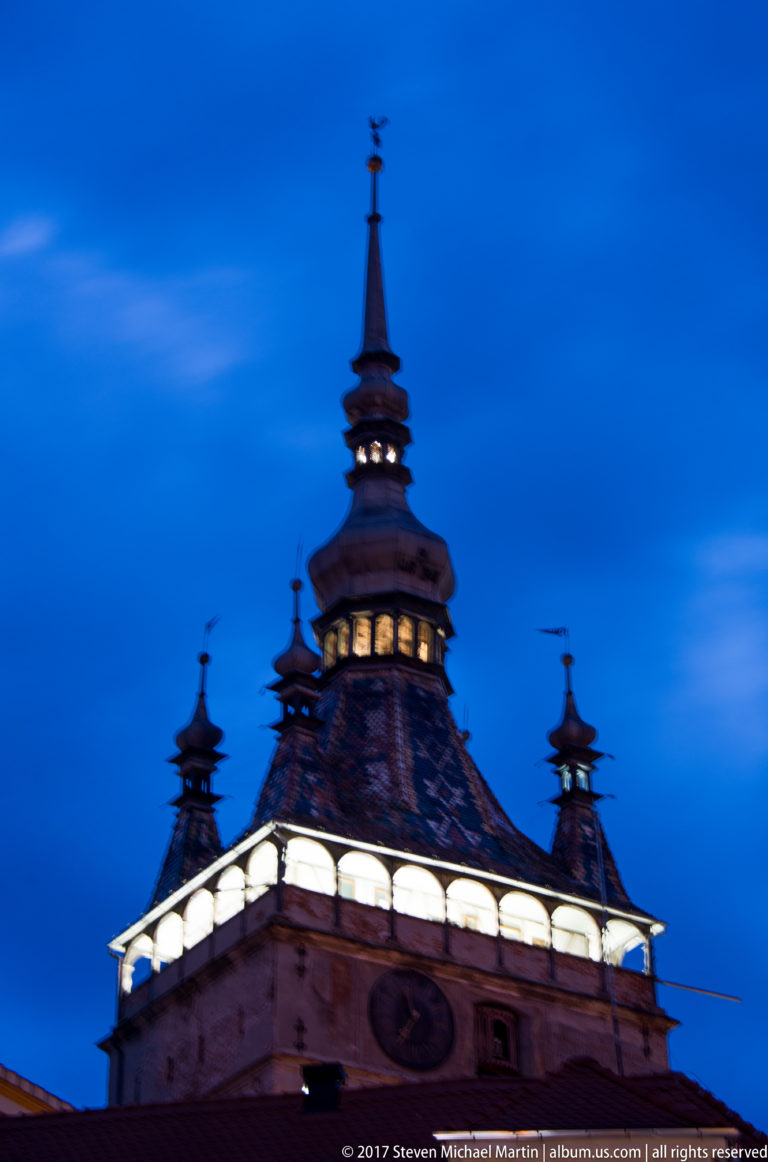 Upper roof, corner turrets, and central spire of Sighisoara Clock Tower against a deep blue sky.