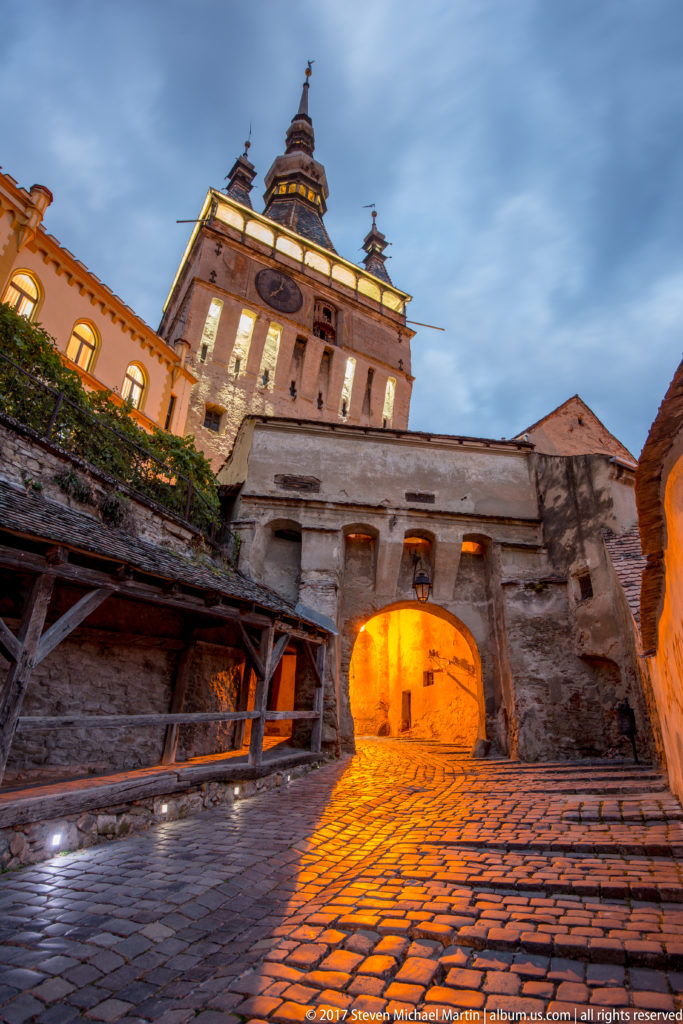 Sighisoara Clock Tower above a lit arched gateway and cobbled approach at dusk.